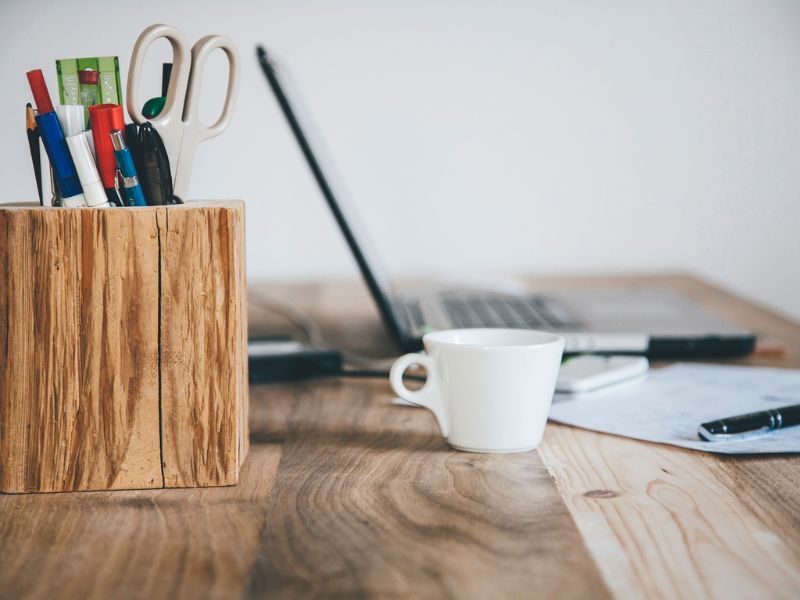 scrivania di legno su cui è appoggiato un porta penne, un pc, una tazzina da caffè e dei fogli di lavoro